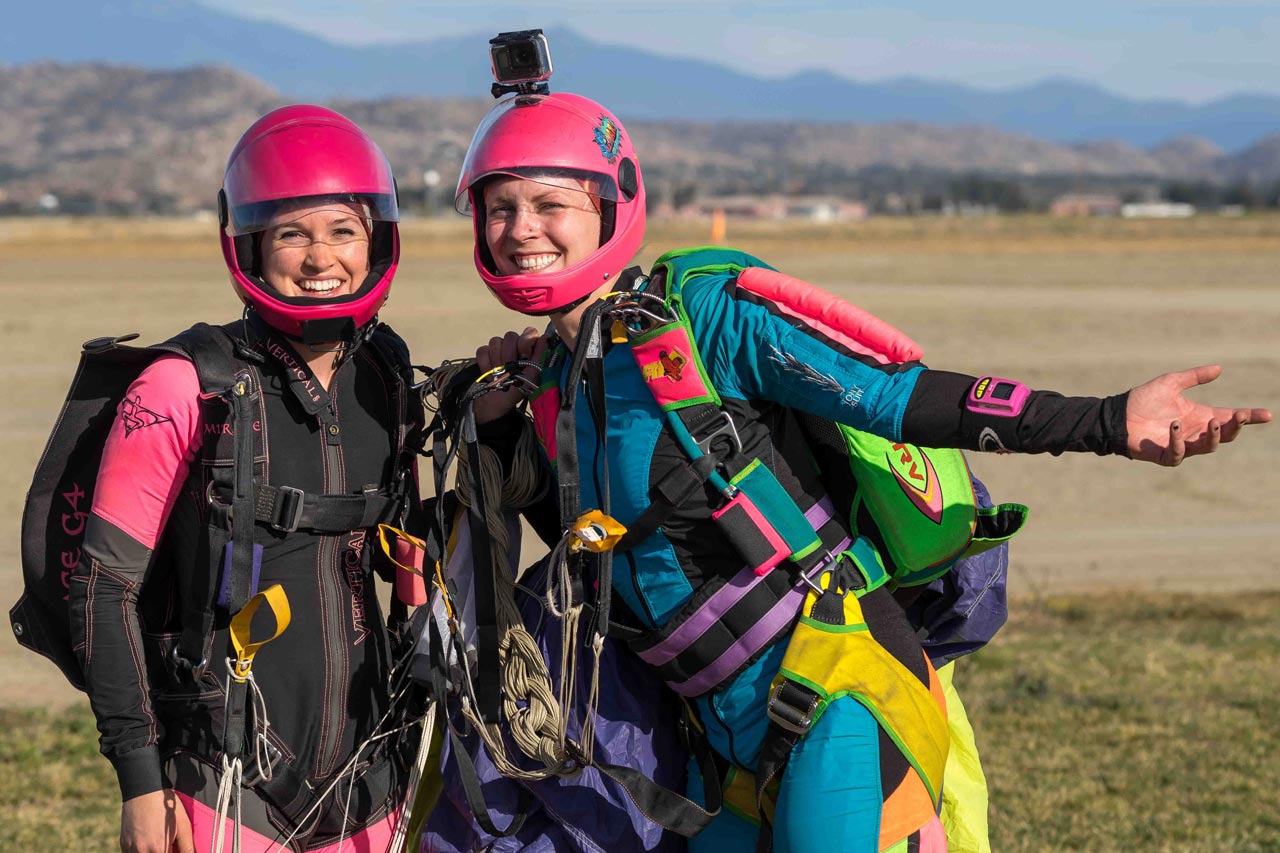 Two female skydivers with pink helmets and colorful rigs stand and pose for the camera