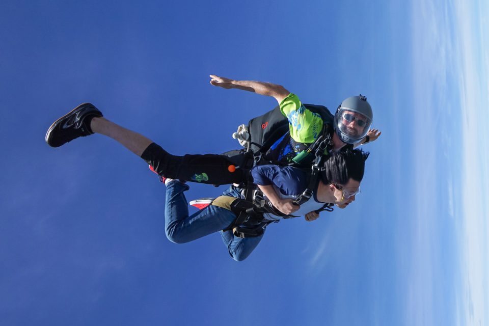 Two people are tandem skydiving, wearing harnesses and gear, free-falling through a clear blue sky. The instructor is positioned above the other person, who has sunglasses and is gripping the harness straps.