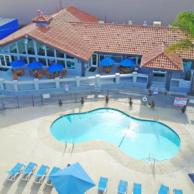 Concrete surrounds a light blue pool in front of a blue building with terra cotta tiles on the roof