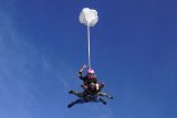 Two people tandem skydiving with an open parachute against a clear blue sky. The person on top wears a pink helmet and makes hand signs, while the instructor below steers the parachute.