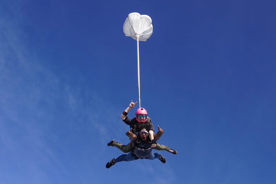Two people tandem skydiving with an open parachute against a clear blue sky. The person on top wears a pink helmet and makes hand signs, while the instructor below steers the parachute.