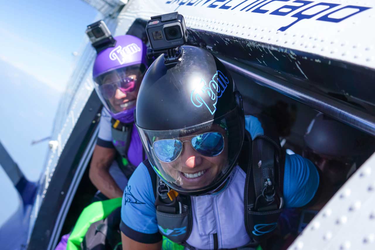 Two skydivers wearing helmets, sunglasses, and harnesses prepare to jump from an airplane. Both have cameras mounted on their helmets and are smiling, with blue sky visible in the background.