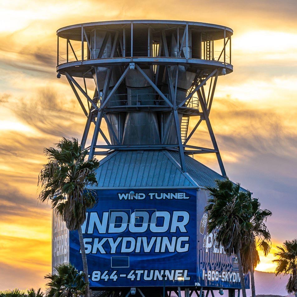 Exterior view of the wind tunnel at Skydive Perris with palm trees in front and sunset with clouds in the background