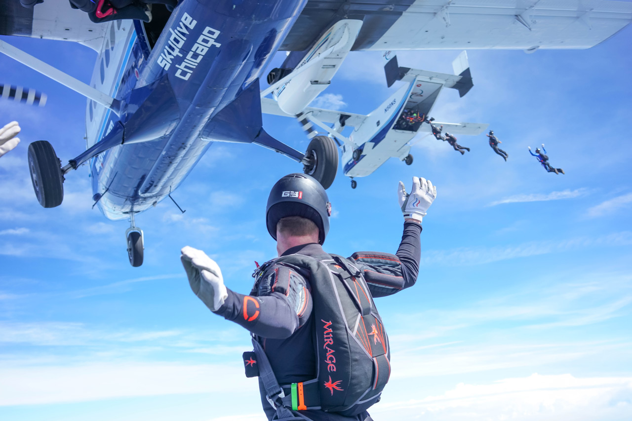 A skydiver with arms raised prepares to jump from a plane, watching other skydivers exit nearby aircraft against a bright blue sky with white clouds. “Skydive Chicago” is visible on the closest plane.
