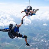 Outside view of a tandem skydiving student and instructor in freefall holding on to the foot of a videographer